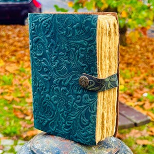 May include: A teal leather-bound journal with a floral embossed design. The journal has a brown leather strap with a metal clasp and is closed.