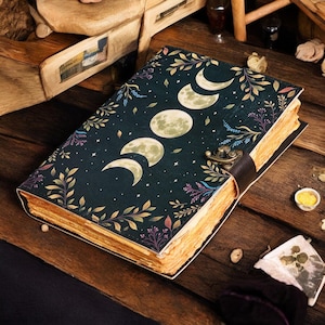 May include: A dark green journal with a celestial moon phase design. The cover features a crescent moon, half-moons, and full moons, surrounded by leaves and stars. The journal has a leather strap closure and aged-looking pages.