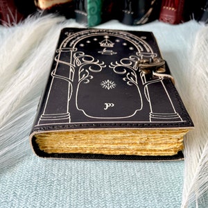 Puede incluir: Un diario de cuero negro con un cierre y un candado plateados. La portada presenta un dise&ntilde;o blanco de un &aacute;rbol con una estrella y una corona encima. El diario tiene una encuadernaci&oacute;n dorada y p&aacute;ginas.