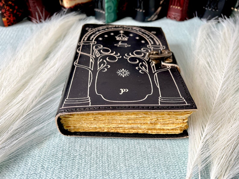 Puede incluir: Un diario de cuero negro con un cierre y un candado plateados. La portada presenta un dise&ntilde;o blanco de un &aacute;rbol con una estrella y una corona encima. El diario tiene una encuadernaci&oacute;n dorada y p&aacute;ginas.