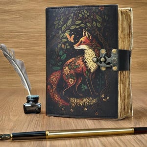 Könnte beinhalten: Ein ledergebundenes Notizbuch mit einer Fuchsgrafik, einer Feder, einem Tintenfass und einem Stift. Das Notizbuch zeigt ein detailliertes Design eines Fuchses in einer Waldszene mit einem Schmetterling. Das Notizbuch ist mit einem Metallverschluss verschlossen.