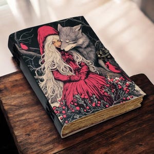 Könnte beinhalten: Ein ledergebundenes Notizbuch mit einer Fantasy-Illustration eines Mädchens in rotem Kleid und Kapuze, das einen Wolf umarmt. Das Cover zeigt einen schwarzen Hintergrund mit roten Beeren und Vögeln. Das Notizbuch hat einen Metallverschluss.