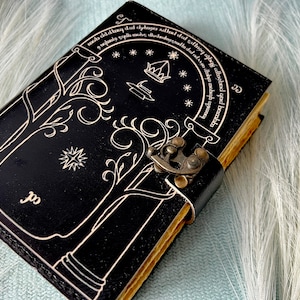 Puede incluir: Un diario de cuero negro con un dise&ntilde;o decorativo de &aacute;rboles y una cerradura. El diario tiene una cita en escritura &eacute;lfica alrededor del borde. La cita dice "mob dddag dul chybager Adhad dad dhoog abge camehed qad bambbs".