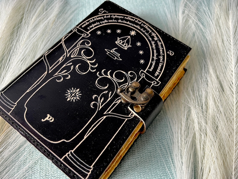 Puede incluir: Un diario de cuero negro con un dise&ntilde;o decorativo de &aacute;rboles y una cerradura. El diario tiene una cita en escritura &eacute;lfica alrededor del borde. La cita dice "mob dddag dul chybager Adhad dad dhoog abge camehed qad bambbs".