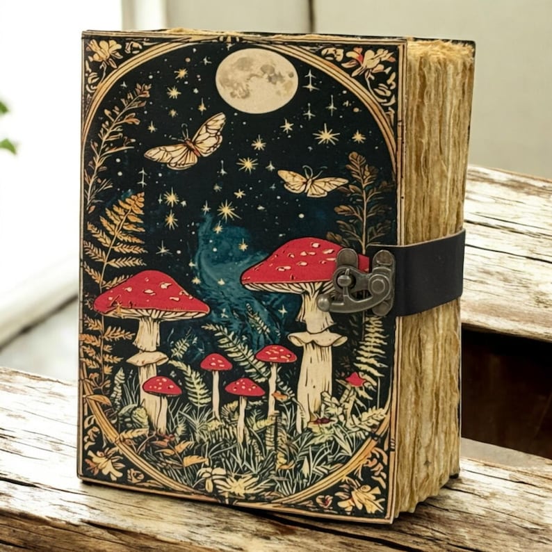 May include: A decorative journal with a whimsical design. The cover features a night scene with red mushrooms, butterflies, and a full moon. The journal has a black leather strap closure and aged paper edges.
