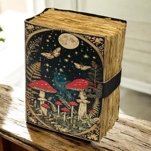 May include: A vintage-style journal with a whimsical design. The cover features a dark background with mushrooms, butterflies, and a full moon. The journal is secured with a black leather strap and a metal clasp. The pages have a rustic, aged appearance.
