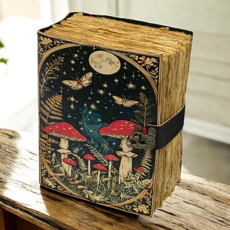 May include: A vintage-style journal with a whimsical design. The cover features a dark background with mushrooms, butterflies, and a full moon. The journal is secured with a black leather strap and a metal clasp. The pages have a rustic, aged appearance.