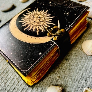 May include: A black leather journal with a gold sun and moon design. The journal has a clasp closure and is decorated with stars. The journal is lying on a grey textured surface.