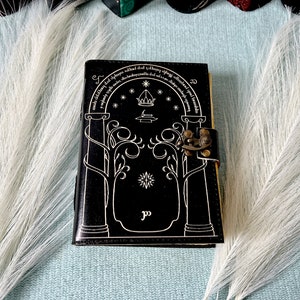 Puede incluir: Diario de cuero negro con un dise&ntilde;o blanco de una puerta con &aacute;rboles y estrellas. El diario tiene un cierre de lat&oacute;n y el texto "Not all those who wander are lost" est&aacute; escrito alrededor de la parte superior de la puerta.