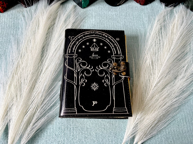 Puede incluir: Diario de cuero negro con un dise&ntilde;o blanco de una puerta con &aacute;rboles y estrellas. El diario tiene un cierre de lat&oacute;n y el texto "Not all those who wander are lost" est&aacute; escrito alrededor de la parte superior de la puerta.