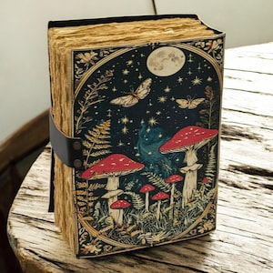 May include: A hardcover journal with a fantasy illustration. The cover features a dark blue night sky with a full moon, butterflies, and red mushrooms. A black leather strap secures the journal.