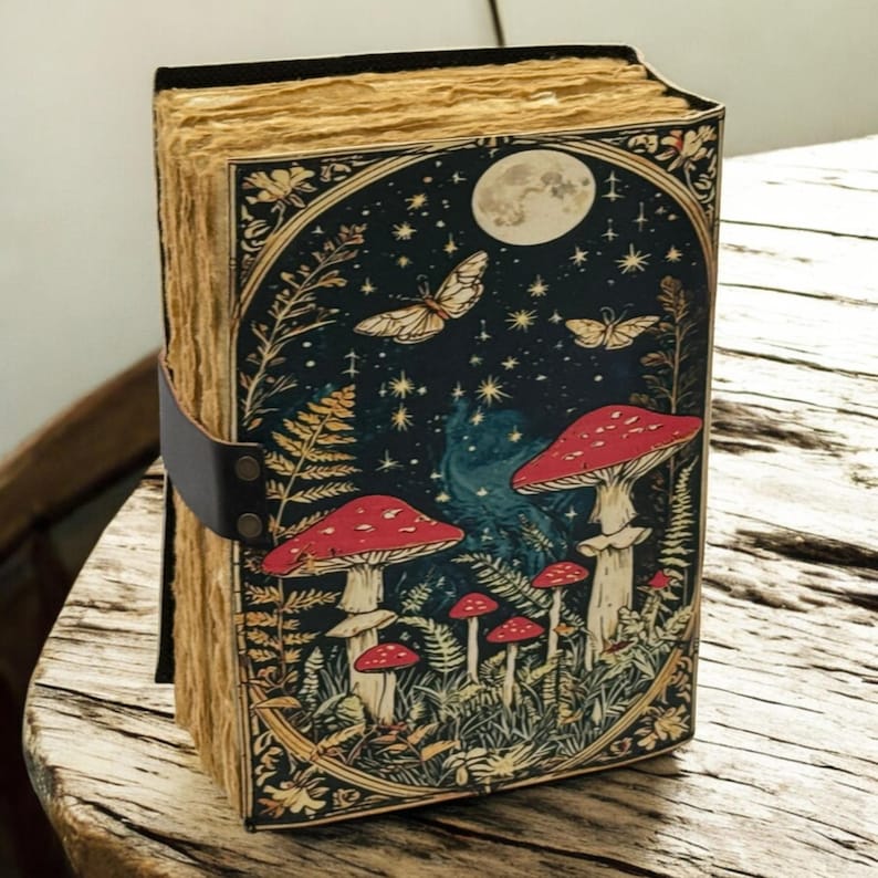 May include: A hardcover journal with a fantasy illustration. The cover features a dark blue night sky with a full moon, butterflies, and red mushrooms. A black leather strap secures the journal.
