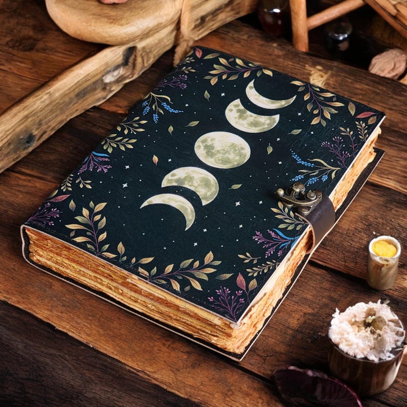 May include: A dark blue journal with a celestial design featuring moon phases and floral accents. The cover is adorned with a decorative clasp and a leather strap. The pages have a vintage, aged appearance.