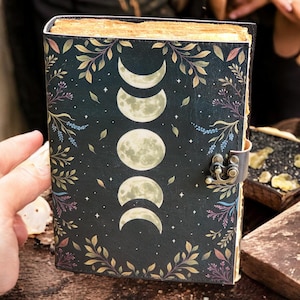 May include: A dark-covered journal with a moon phase design. The cover features a series of moon phases, surrounded by botanical illustrations and stars. The journal has a metal clasp and aged-looking pages.