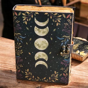 May include: A dark-covered journal with a moon phase design. The cover features a black background with a crescent moon, half-moon, and full moon illustration. The journal is secured with a metal clasp and leather strap, and is surrounded by floral and leaf accents.