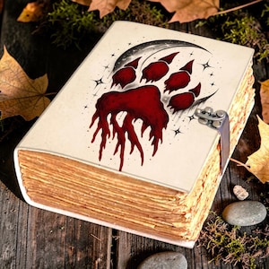 Peut inclure: Un livre rectangulaire à couverture crème orné d'un motif de patte rouge foncé dégoulinante, centré dans un croissant de lune noir avec des étoiles. Le livre possède une fermeture à loquet métallique et des pages épaisses en papier beige.