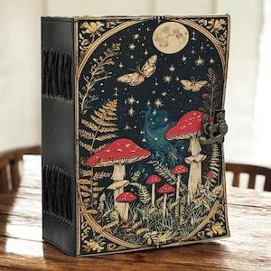 May include: A black leather-bound journal with a fantasy illustration on the front cover. The cover features a night scene with red mushrooms, butterflies, a full moon, and stars. The journal has a metal clasp and decorative stitching.
