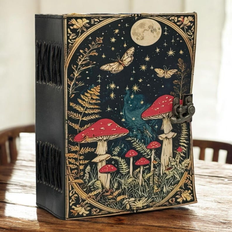 May include: A black leather-bound journal with a fantasy illustration on the front cover. The cover features a night scene with red mushrooms, butterflies, a full moon, and stars. The journal has a metal clasp and decorative stitching.