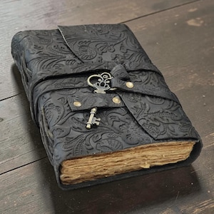 May include: A black journal with an embossed floral pattern on the cover, featuring a wrap-around strap secured by a metallic key-shaped clasp and two brass-colored rivets. The book contains thick, deckled-edge pages with a light tan, aged appearance.