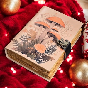 May include: A beige journal with a mushroom illustration and a metal clasp. The cover features orange mushrooms, green leaves, and a peach-colored rose. The journal is closed with a black leather strap and a metal clasp.