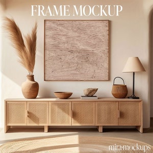 May include: A light wood cabinet with four doors and a light brown rug in front of it. A framed abstract art print with a pink background hangs on the wall above the cabinet. A vase with dried pampas grass and a small bowl are on top of the cabinet. A lamp with a beige lampshade and a woven basket are on the right side of the cabinet. The text "FRAME MOCKUP" is written in white letters on the wall above the art print.