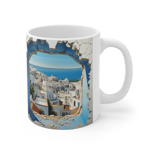 May include: White ceramic coffee mug featuring a scenic view of a coastal town through a broken wall. The image shows white buildings with red roofs, blue sky, and sea. The mug has a white handle.
