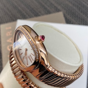May include: A rose gold Bvlgari watch with a serpentine-inspired flexible band. The oval-shaped watch face features a silver sunburst dial with Roman numeral markers and a diamond-encrusted bezel. A small pink cabochon stone is set on the crown.