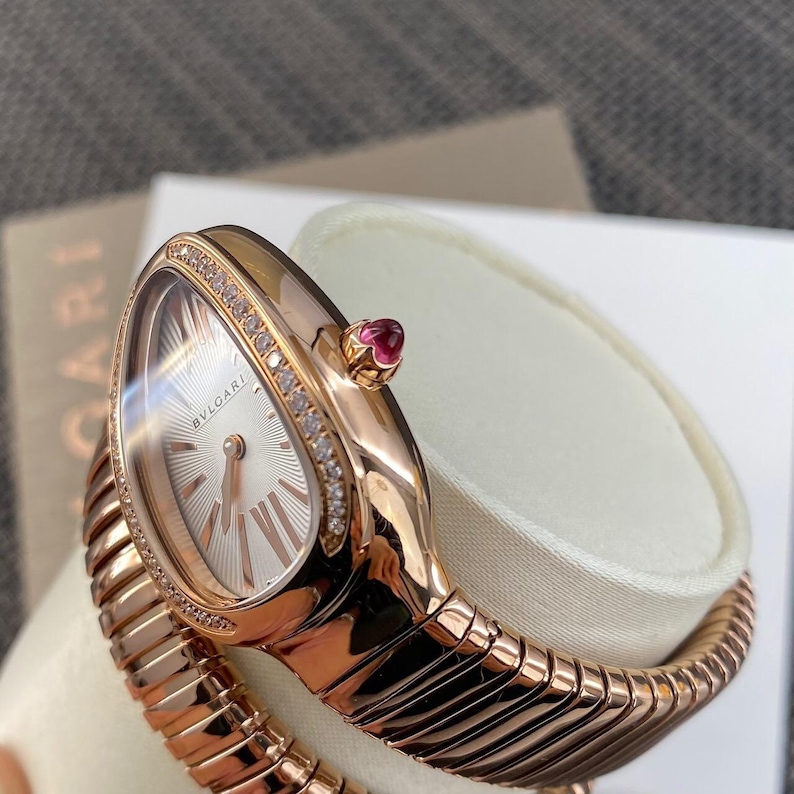 May include: A rose gold Bvlgari watch with a serpentine-inspired flexible band. The oval-shaped watch face features a silver sunburst dial with Roman numeral markers and a diamond-encrusted bezel. A small pink cabochon stone is set on the crown.