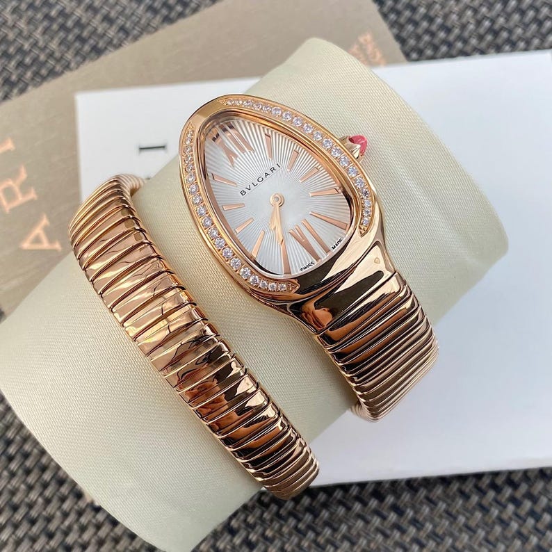 May include: A rose gold Bvlgari watch with a teardrop-shaped case, a white sunburst dial, and a flexible tubogas-style bracelet. The bezel is set with small round diamonds, and the crown features a pink stone accent. The dial displays the Bvlgari logo.