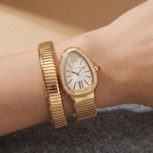 May include: A gold-tone watch with a teardrop-shaped case and a flexible, multi-coil bracelet. The white dial features Roman numeral indices and the BVLGARI logo. The bezel is set with small clear stones, and a small red cabochon is on the crown.