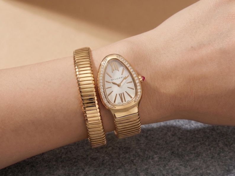 May include: A gold-tone watch with a teardrop-shaped case and a flexible, multi-coil bracelet. The white dial features Roman numeral indices and the BVLGARI logo. The bezel is set with small clear stones, and a small red cabochon is on the crown.