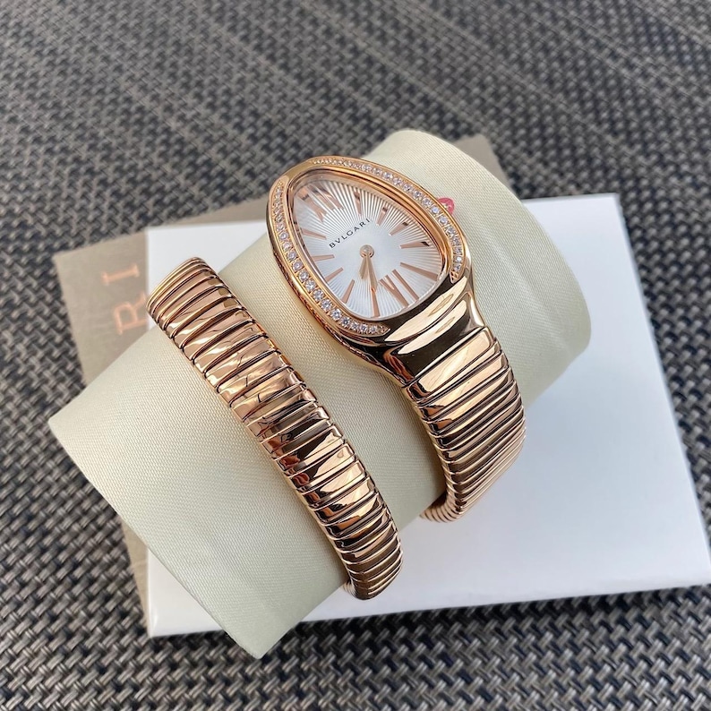 May include: A rose gold Bvlgari Serpenti watch with a teardrop-shaped case, a white dial featuring the Bvlgari logo, and a diamond-set bezel. The watch has a flexible, coiled tubogas-style bracelet that wraps around the wrist in a double loop.