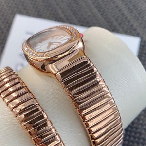 May include: A rose gold-tone wristwatch featuring a curved, rectangular case with a diamond-encrusted bezel. The watch has a flexible, segmented metal bracelet with a polished finish. The white dial includes simple hour markers and hands.