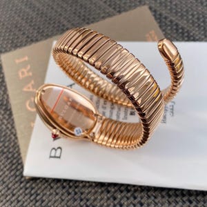 May include: A rose gold-toned watch with a flexible, coiled snake-like band. The watch case is oval-shaped with the text BVLGARI visible on the back. A small red accent is present on the side of the case, and the band features a segmented design.