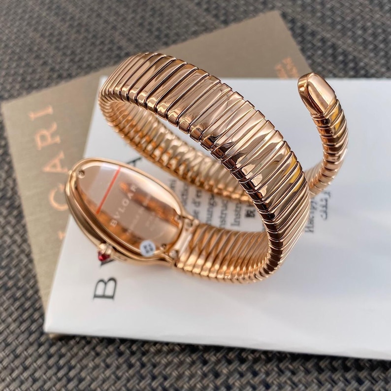 May include: A rose gold-toned watch with a flexible, coiled snake-like band. The watch case is oval-shaped with the text BVLGARI visible on the back. A small red accent is present on the side of the case, and the band features a segmented design.