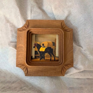 Puede incluir: Pequeño marco cuadrado de madera con una pintura detallada de un carruaje tirado por caballos. El caballo negro tiene una manta amarilla. El marco tiene un borde decorativo tallado, sobre fondo blanco.