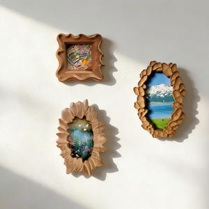 Op de afbeelding: Drie decoratieve houten frames met kunstwerken met een natuurthema hangen aan een witte muur. De frames hebben unieke vormen, waaronder een vierkant, een ovaal en een zonnebloemontwerp. De kunstwerken tonen landschappen en bloemstukken.