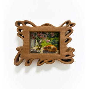 May include: A rectangular wooden picture frame with an organic, cutout border design. The frame holds a small painting depicting an orange cat sleeping on a surface next to a glass vase filled with pink flowers and green foliage in sunlight.