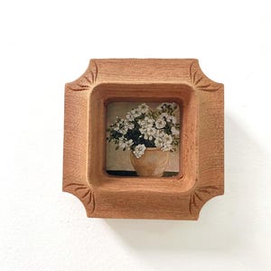 May include: A small, square, wooden frame with a carved design. The frame holds a miniature painting of white flowers in a tan pot. The frame is a warm, light brown colour and is set against a white background.
