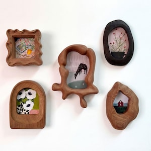 Può includere: Una collezione di cinque cornici in legno uniche, ognuna con una forma organica distinta. Le cornici contengono varie piccole opere d'arte, tra cui motivi floreali, una giraffa e una casa rossa. Le cornici sono montate su un muro bianco.