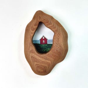 Peut inclure: Une œuvre d'art murale en bois de forme organique. Le cadre entoure une petite peinture d'une maison rouge sur une colline verte, avec un océan et un ciel bleus en arrière-plan. Le cadre mesure environ 30 cm de large.