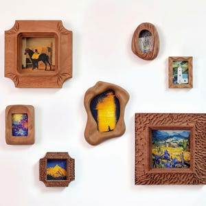 May include: A collection of framed art pieces in various shapes and sizes. The frames are made of wood and contain different landscape and animal scenes. The art pieces are mounted on a white wall.