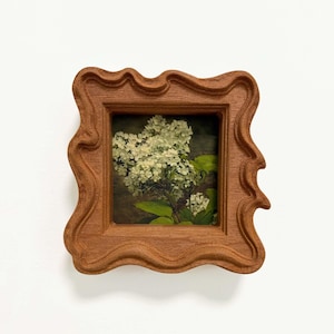 Puede incluir: Un pequeño marco de madera cuadrado con un borde ondulado y orgánico que rodea una imagen impresa de flores de hortensia blancas con hojas verdes. El marco tiene una textura de veta de madera marrón natural y un acabado mate.