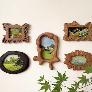 Peut inclure: Cinq cadres photo en bois de formes originales, affichant des paysages. Les cadres, de couleur marron, sont accrochés sur un mur blanc. Les images présentent des scènes de nature avec des verts et des bleus, pour une ambiance champêtre.