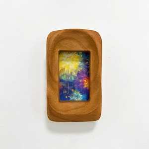 May include: A rectangular wooden frame with a smooth, carved, light brown finish. The center opening displays a vibrant, colorful print featuring yellow, purple, and blue abstract firework-like patterns against a dark blue background.