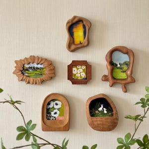 May include: A collection of six wooden framed art pieces, each with a unique organic shape. The artwork within the frames depicts various scenes, including landscapes, floral arrangements, and a pair of rabbits. The frames are mounted on a neutral-colored wall.