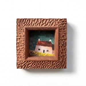 May include: A small, square, wooden frame with a carved, textured surface surrounds a miniature painting of a house. The house has a red roof, white walls, and a black door. The background is green.