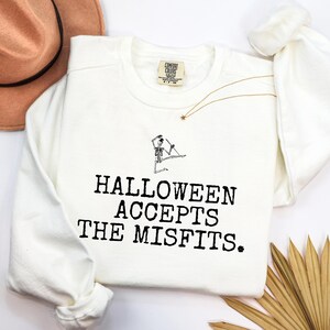 May include: An off-white long-sleeve crewneck sweatshirt featuring a black graphic of a skeleton marionette and the text "HALLOWEEN ACCEPTS THE MISFITS." The garment has a Comfort Colors brand label at the inner neckline and long sleeves.