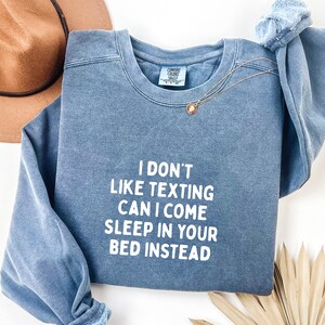 May include: A blue crewneck sweatshirt with the text "I DON'T LIKE TEXTING CAN I COME SLEEP IN YOUR BED INSTEAD" in white. A gold necklace with a pendant is draped on the sweatshirt. A brown hat is visible in the background.