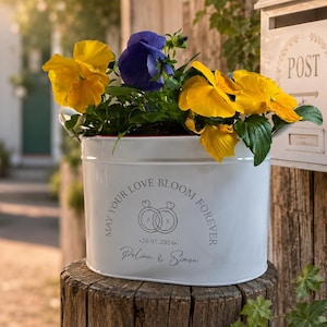 Könnte beinhalten: Ein weißer, ovaler Metallübertopf gefüllt mit gelben und violetten Stiefmütterchen. Die Vorderseite zeigt den Text "MAY YOUR LOVE BLOOM FOREVER", eine Abbildung von zwei ineinander verschlungenen Ringen, das Datum "20.07.2024" und die Namen "Polina & Simon" in Grau.